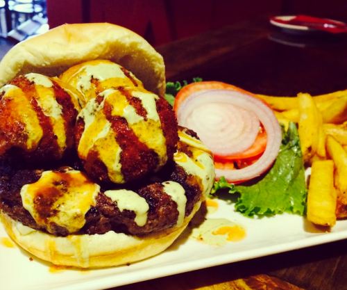 A burger with three crispy patties, drizzled with sauce, served with lettuce, tomato, and fries on the side.