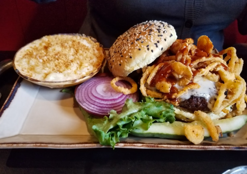 A plate featuring a burger with onion rings, lettuce, tomato, and a side of creamy mac and cheese.