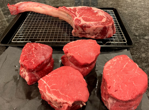 A large ribeye steak with a long bone and several smaller beef tenderloin cuts on a wire rack and black paper.