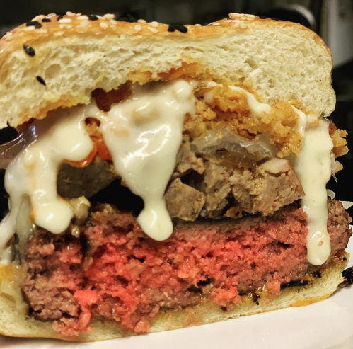 Close-up of a layered burger with pink beef patty, crispy toppings, and creamy sauce in a sesame seed bun.