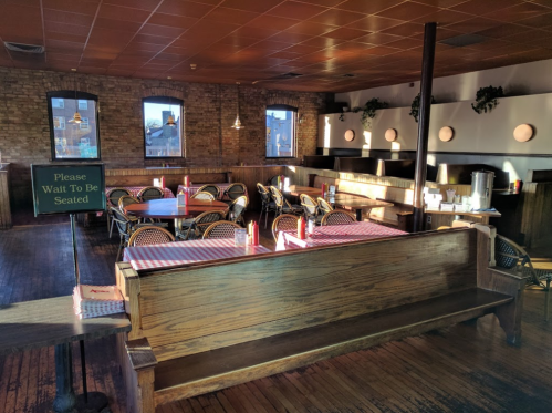 A cozy restaurant interior with wooden seating, checkered tablecloths, and a "Please Wait To Be Seated" sign.