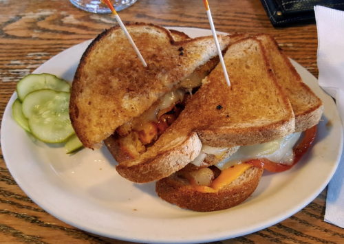 A toasted sandwich with melted cheese, grilled onions, and pickles on the side, served on a white plate.