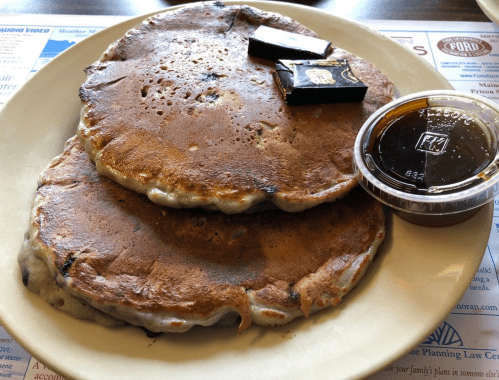 Two large, golden-brown pancakes topped with chocolate squares and served with a side of syrup.