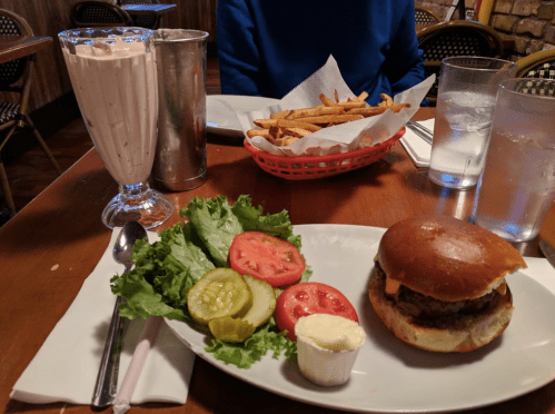 A burger with lettuce, tomato, pickles, and mayo, served with fries and a milkshake in a diner setting.