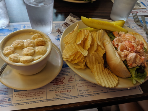 A bowl of clam chowder, a lobster roll with lettuce, potato chips, and a pickle on a plate.