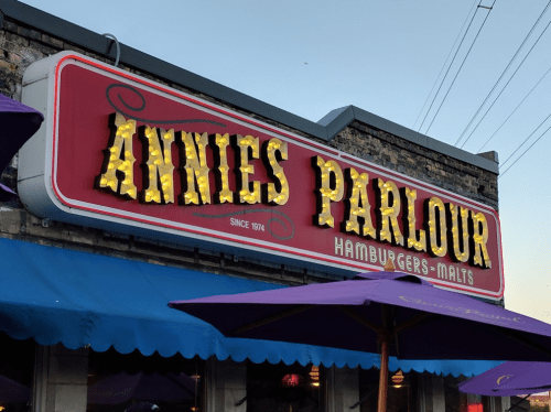 Sign for Annie's Parlour, featuring bright lights and the text "Hamburgers-Malts," with outdoor seating below.
