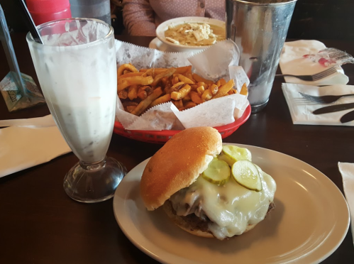 A cheeseburger with pickles on a plate, accompanied by a glass of milkshake and a basket of fries in the background.
