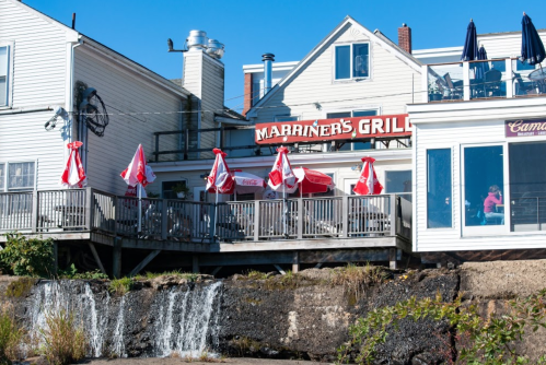 A waterfront restaurant with red umbrellas on the deck, featuring a sign that reads "Mariner's Grill" and a small waterfall.