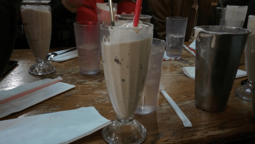 Milkshakes in tall glasses with straws, surrounded by empty glasses and napkins on a wooden table.