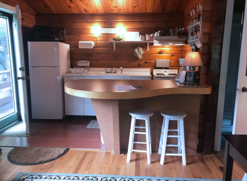 Cozy kitchen with wooden walls, a curved countertop, white appliances, and two stools. Bright and inviting atmosphere.