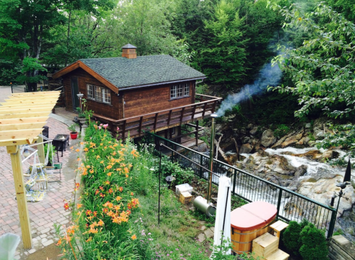 A wooden cabin by a stream, surrounded by greenery and flowers, with smoke rising from a chimney.