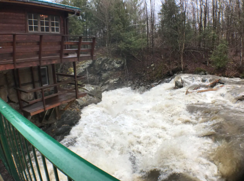 A rushing river flows beside a wooden cabin, surrounded by trees, with a green railing in the foreground.