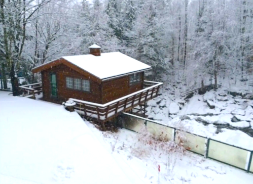 A cozy wooden cabin surrounded by snow-covered trees and a frozen stream, creating a serene winter scene.