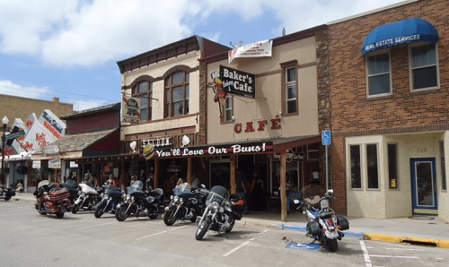 A street view of Baker's Café with parked motorcycles in front and colorful storefronts under a partly cloudy sky.