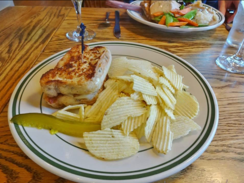 A plate with a grilled sandwich, potato chips, a pickle, and a glass of water in the background.