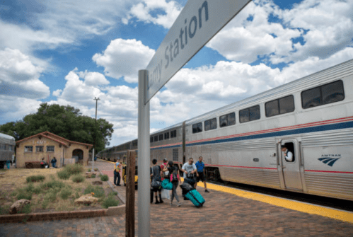 Passengers with luggage board an Amtrak train at a station under a cloudy sky. A building is visible in the background.