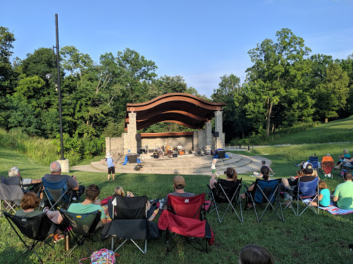 A crowd sits on lawn chairs watching a live performance at an outdoor stage surrounded by trees.