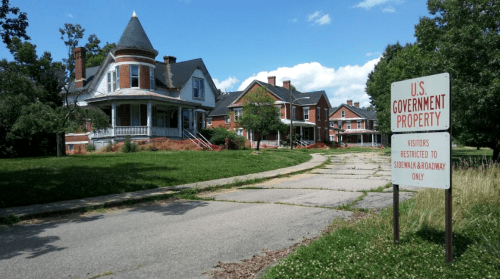 Abandoned houses with a "U.S. Government Property" sign, surrounded by overgrown grass and trees under a blue sky.