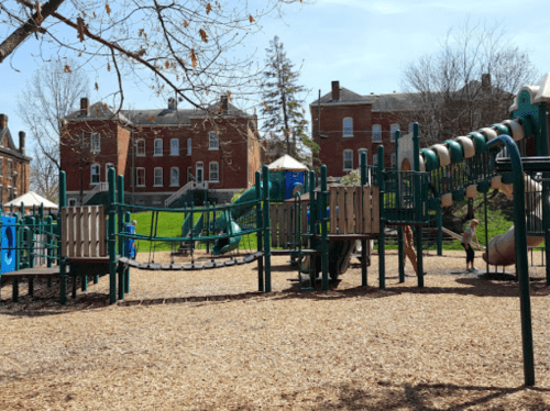 A playground with slides and climbing structures, surrounded by trees and brick buildings in the background.