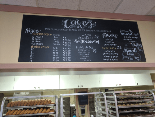 Chalkboard menu displaying cake sizes, prices, fillings, and decorations at a bakery.