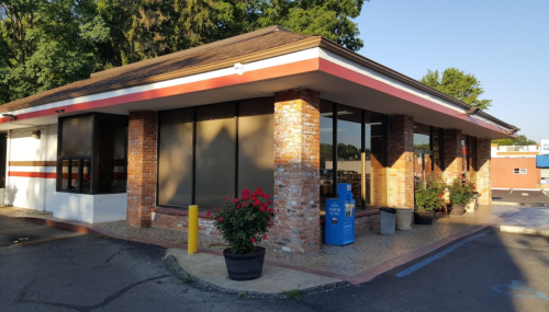 A brick building with large windows, surrounded by greenery and flower pots, featuring a drive-thru area.