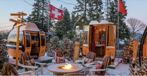 Cozy winter scene with snow-covered cabins, flags, and a fire pit surrounded by festive decorations and trees.
