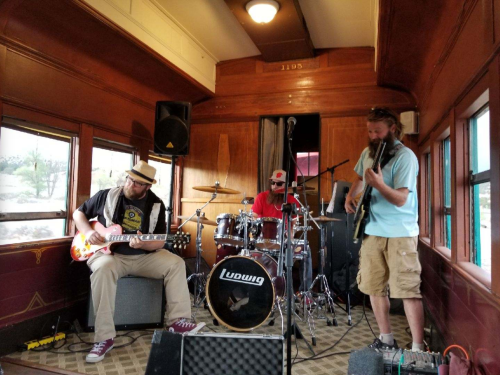 A band performs inside a vintage train car, featuring a guitarist, drummer, and bassist.