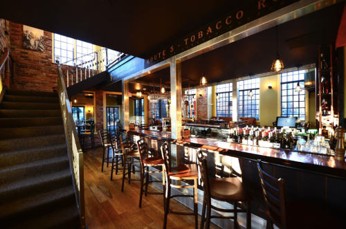 A stylish bar interior with wooden floors, high ceilings, and large windows, featuring a staircase and a well-stocked counter.