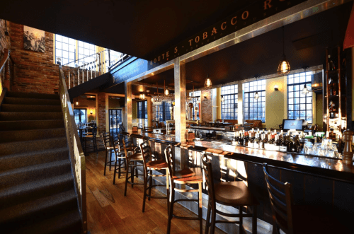 A stylish bar interior with wooden floors, high ceilings, and large windows, featuring a staircase and a well-stocked counter.