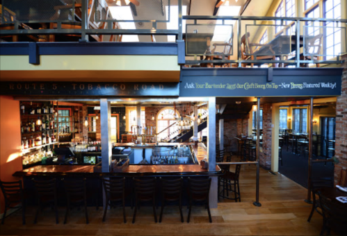 A spacious restaurant interior featuring a bar, wooden chairs, and a two-level seating area with large windows.