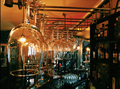 A bar scene featuring hanging glassware and shelves stocked with various bottles, illuminated by warm lighting.
