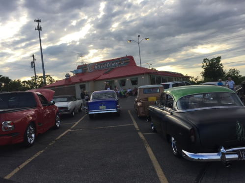 A row of classic cars parked outside a diner under a cloudy sky, with people gathered nearby.