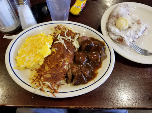 A plate of breakfast with scrambled eggs, hash browns, meatloaf with gravy, and a powdered sugar beignet on the side.