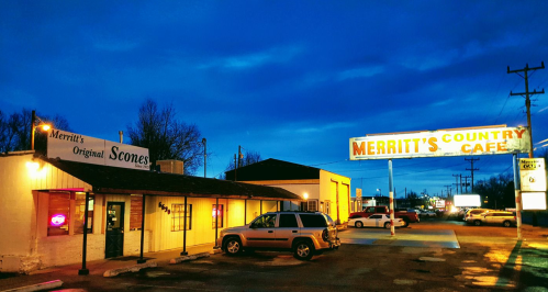 A cozy diner and bakery at dusk, featuring a sign for Merritt's Country Café and a glowing "Scones" sign.