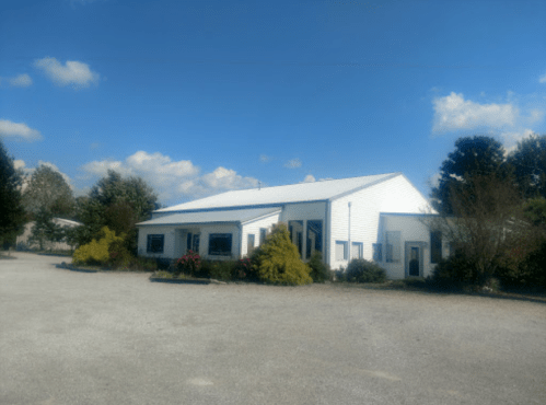 A white building with a metal roof surrounded by greenery and blue skies, set in a gravel parking area.