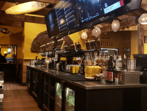 A bar with multiple beer taps, shelves of liquor, and two large screens displaying information in a warm, wooden setting.