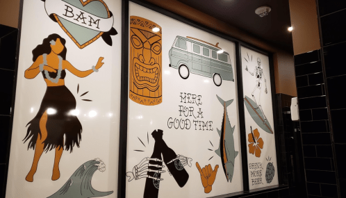 Colorful mural featuring a hula dancer, tiki mask, van, skeleton, and playful phrases about having a good time.
