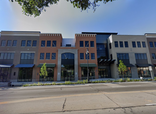 Modern multi-story building with a mix of brick and glass, featuring large windows and greenery in front.