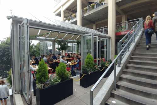 A glass-enclosed restaurant with diners, surrounded by greenery, next to a staircase in an urban setting.