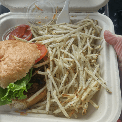A hand holding a takeout container with a sandwich and a side of crispy fries, accompanied by a small cup of ketchup.
