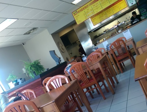 A casual restaurant interior with wooden tables, chairs, and a counter in the background. A staff member is visible.