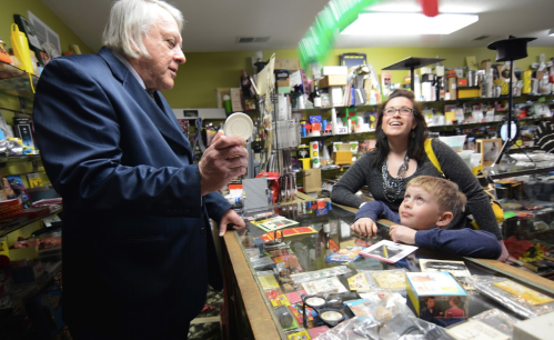 A man in a suit shows a small item to a woman and a child in a colorful shop filled with various products.