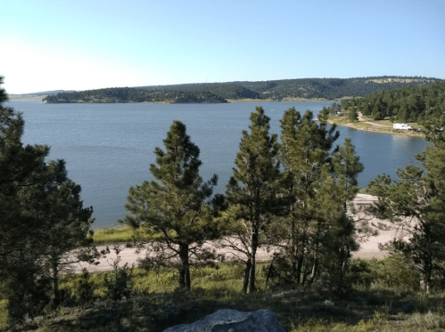 A serene lake surrounded by trees and hills under a clear blue sky. A road runs along the water's edge.