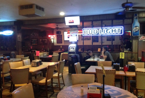 A dimly lit bar with empty tables, neon signs, and two patrons sitting at a table, enjoying their drinks.