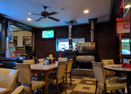 Cozy restaurant interior with wooden tables, chairs, and a large smoker in the background. Warm lighting and decor.