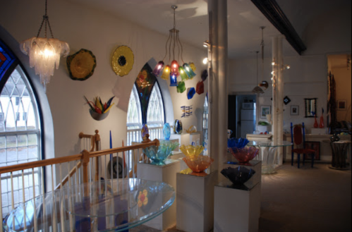 A brightly lit gallery showcasing colorful glass art pieces displayed on shelves and walls.