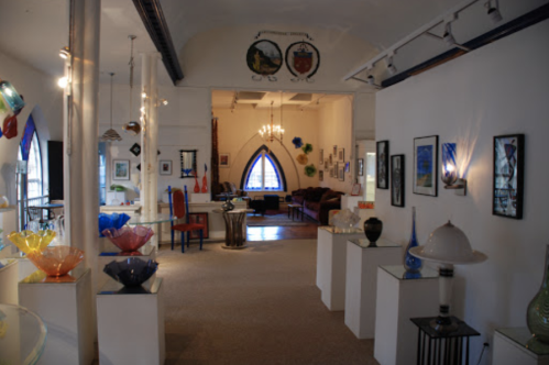 A bright art gallery featuring colorful glass sculptures, paintings, and a cozy seating area in the background.