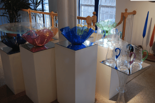 Colorful glass bowls and vases displayed on white pedestals in a bright gallery setting.