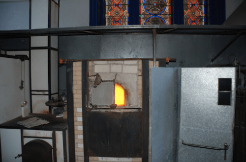 Interior view of a room with a furnace glowing orange, a metal door, and stained glass windows above.