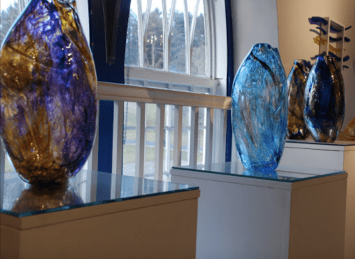 Three colorful glass vases in blue and gold displayed on pedestals near a window.